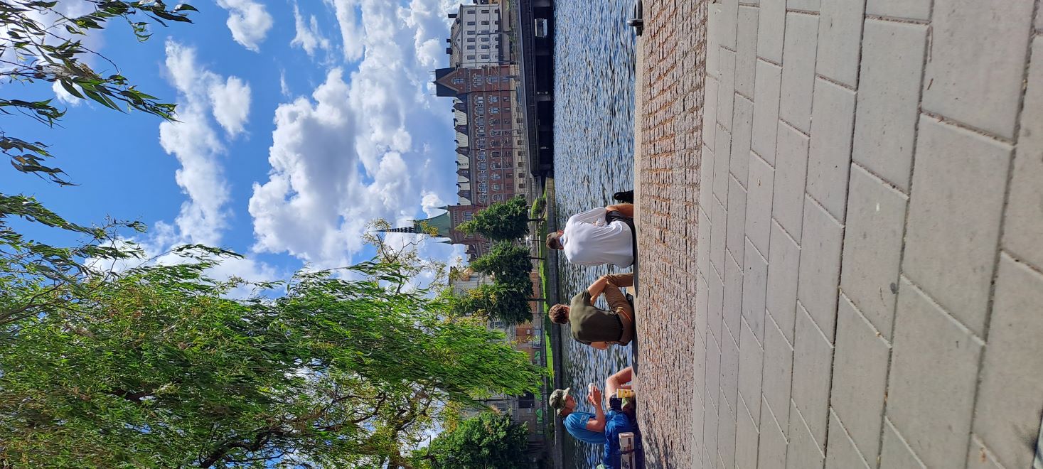 Three boys are seated by the water in Stockholm on a sunny day, enjoying a meal from McDonald's. The photo is taken from behind.