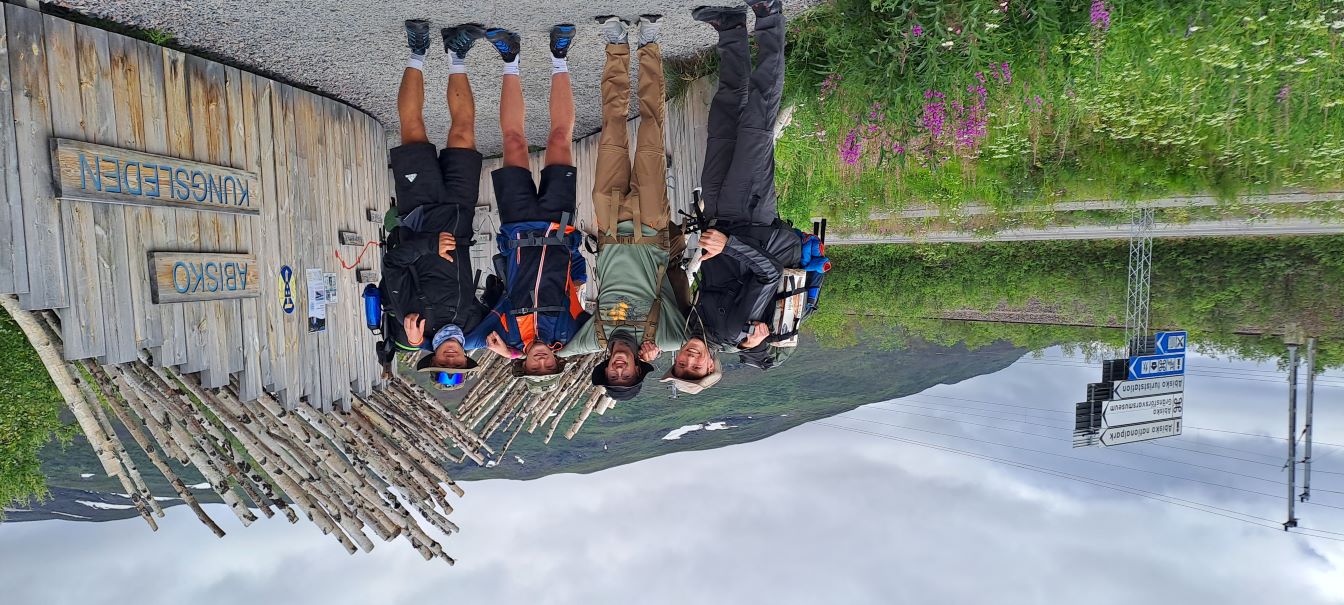 A group of four young men—Florian, Maxi, Niki, and Jonas—in full hiking gear, standing in front of the gate for the Kungsleden trail in Sweden. They appear ready to begin their week-long trek.