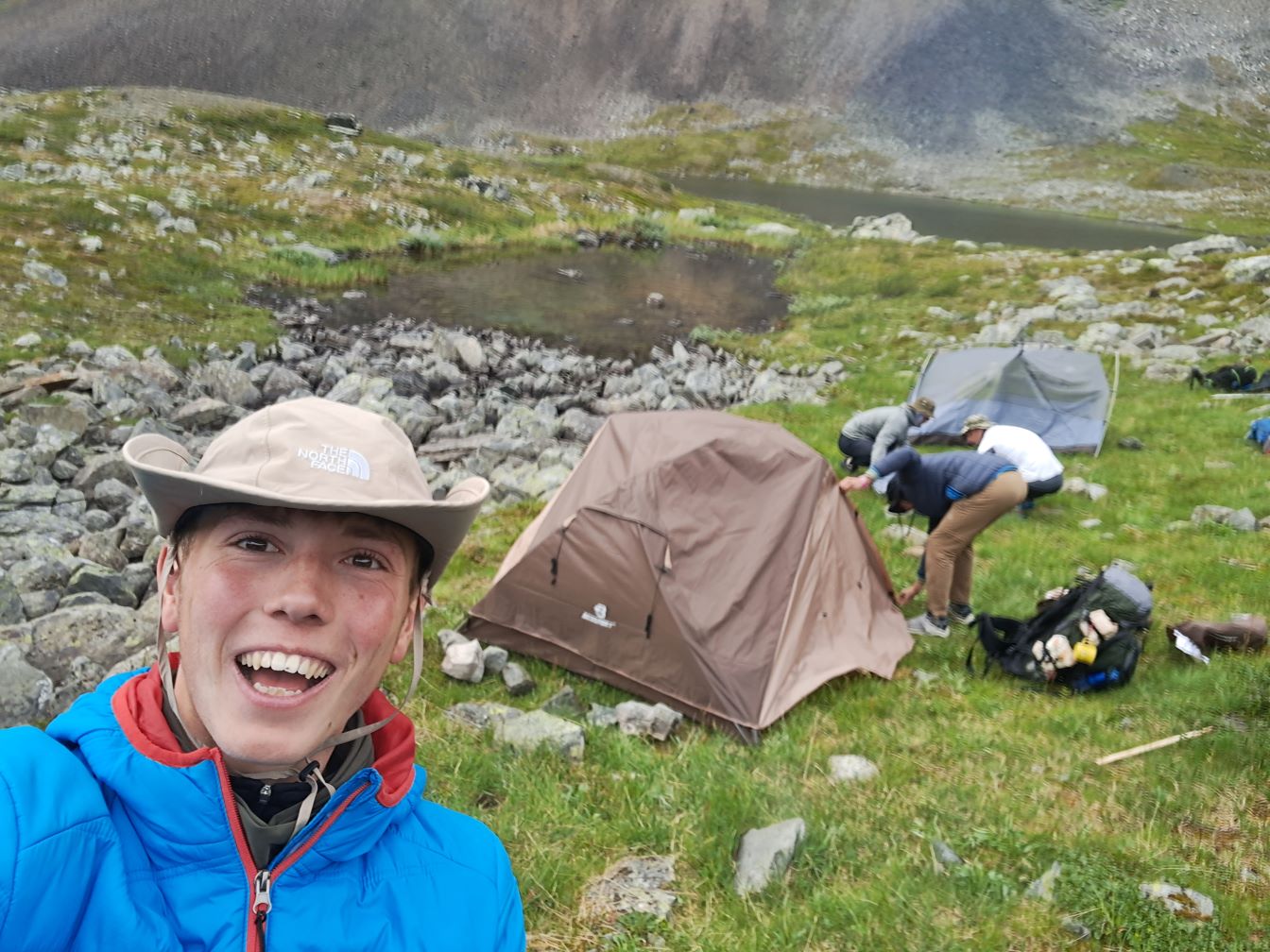 A selfie of Maxi wearing a hat, with the other boys visible in the background setting up tents in a remote location along the Kungsleden trail.